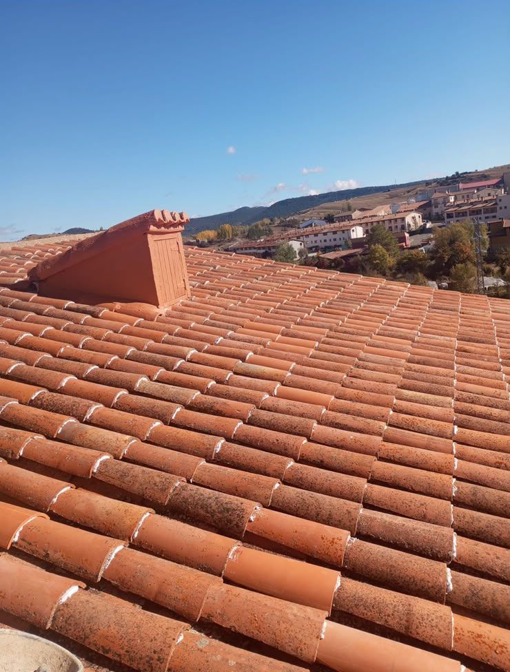 Tejado de tejas de terracota con vistas a un pueblo en la ladera de una colina bajo un cielo azul despejado.