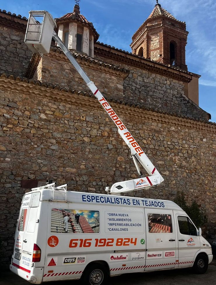 Camión utilitario blanco con plataforma elevadora trabajando en el muro de piedra de una iglesia bajo un cielo azul.