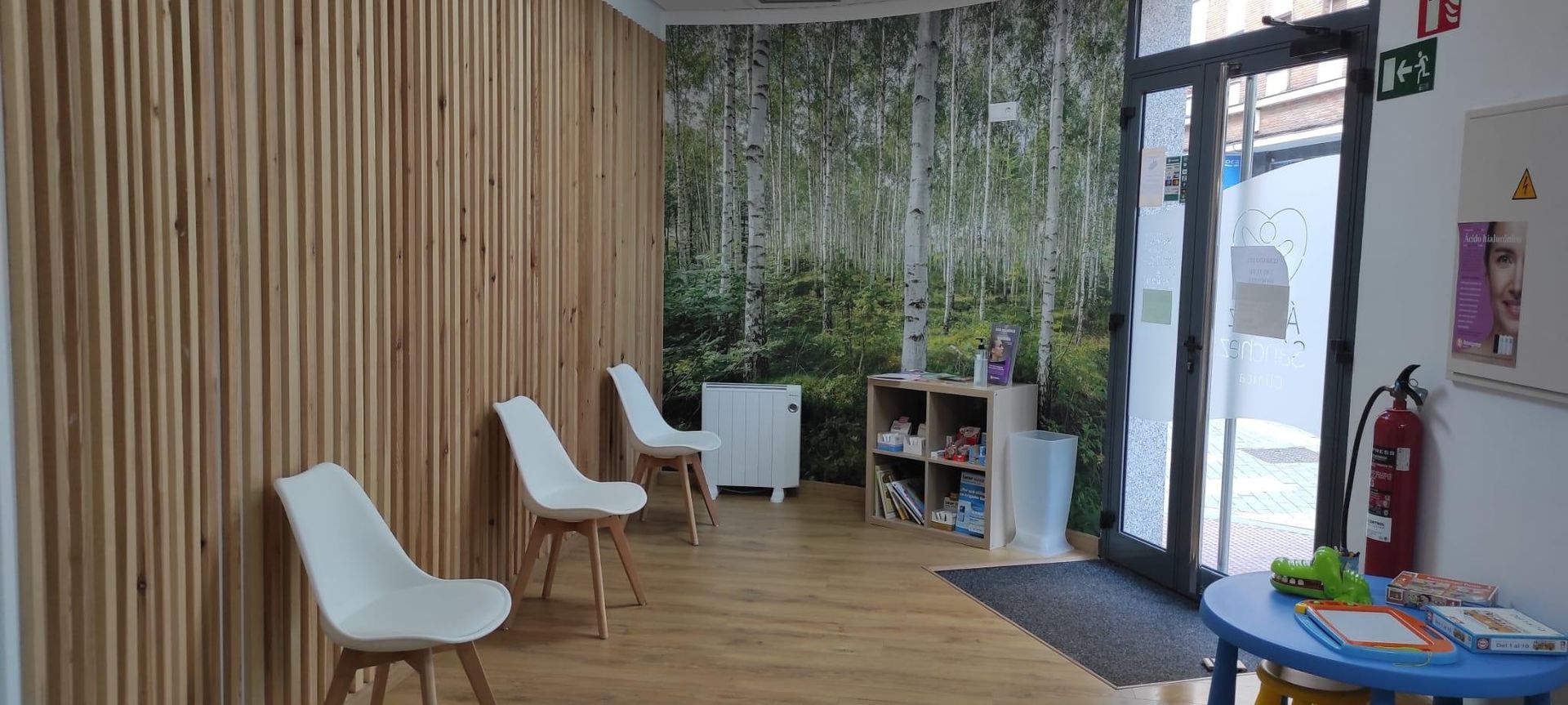 Sala de espera con paneles de madera, sillas blancas y un mural de un bosque. Hay un extintor y una mesa con juguetes.