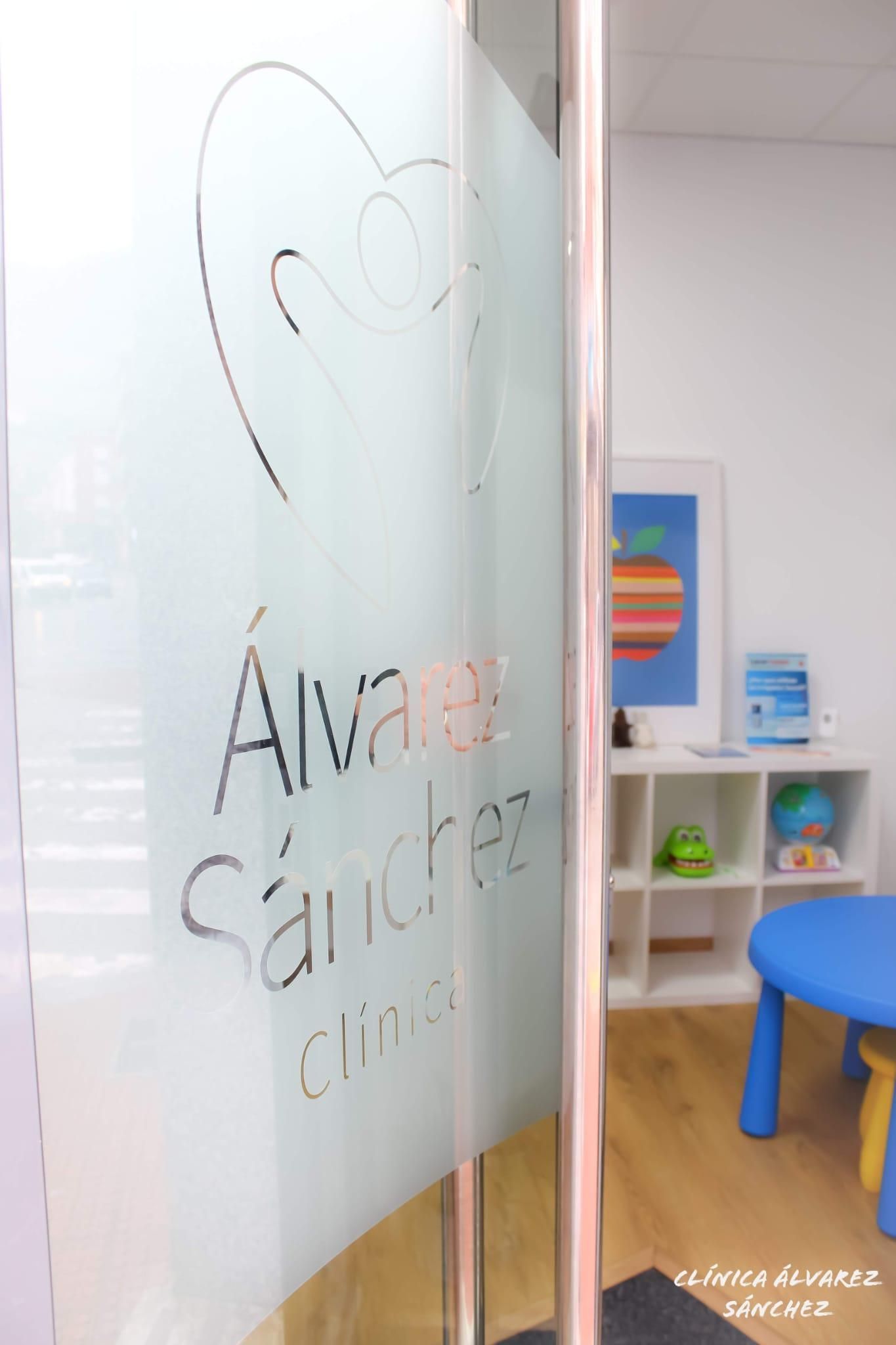Puerta de vidrio con logo de “Álvarez Sánchez Clínica”, zona infantil visible: mesa azul, estantes blancos con juguetes, cuadros coloridos enmarcados.