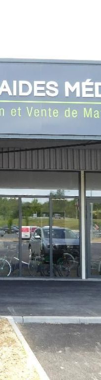Bâtiment avec des vitrines reflétant une voiture, enseigne indiquant AIDES MÉDICALES.