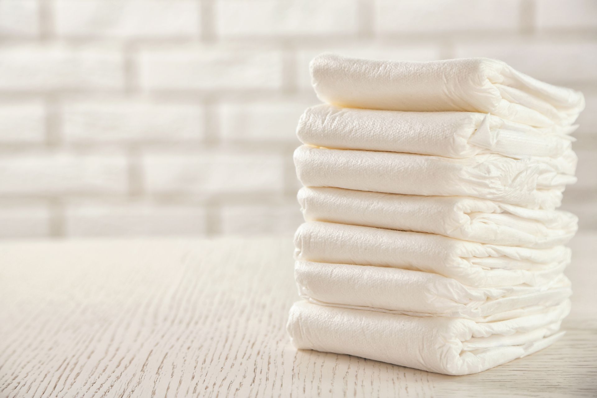 Pile de couches blanches pour bébés sur une surface en bois blanc, avec un mur de briques en arrière-plan.