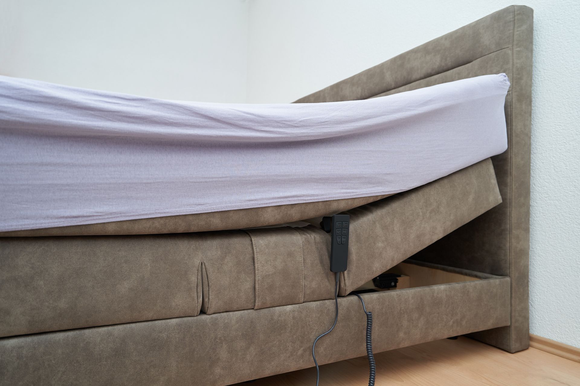 Lit réglable avec tête de lit relevée, recouvert d'un drap gris. Tête de lit et sommier marron.