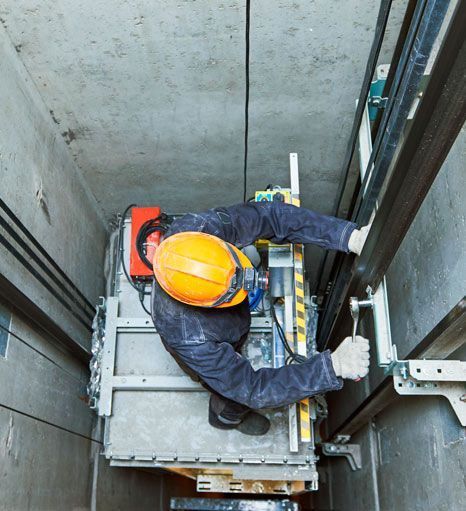 Persona con casco reparando la maquinaria del ascensor dentro de un hueco.