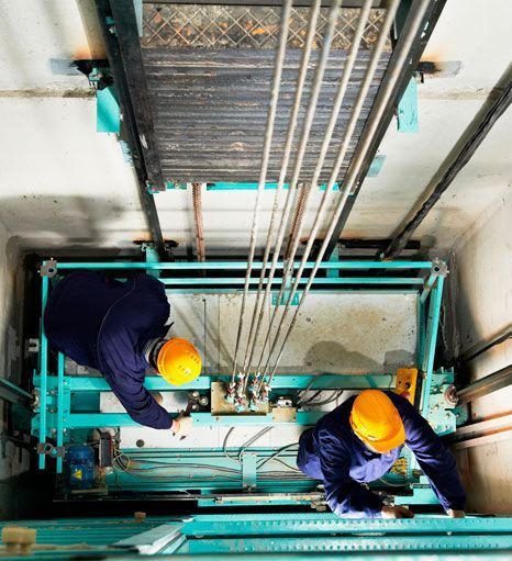 Dos técnicos con monos azules y cascos amarillos inspeccionan los cables del ascensor en un hueco.