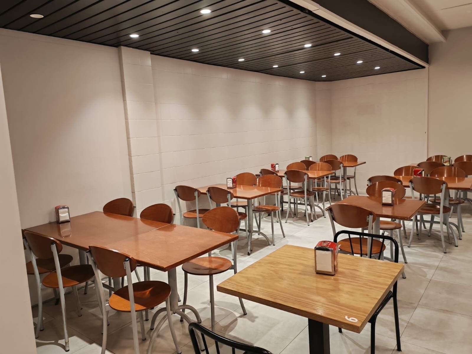 Área de comedor de restaurante vacía con mesas de madera, sillas e iluminación empotrada.