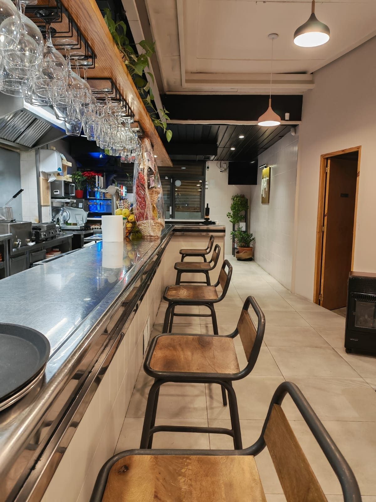 Vista interior de una barra con taburetes, mostrador y cocina visibles.