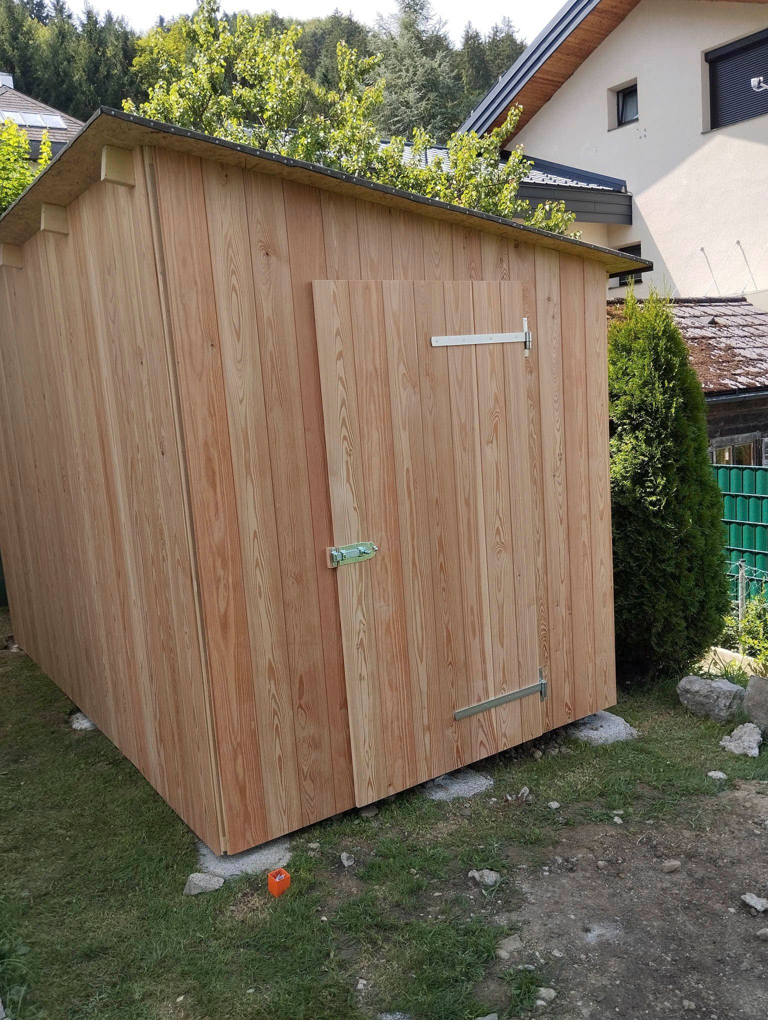 Vor einem Haus steht im Gras ein Holzschuppen.