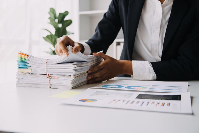 Una persona con traje clasifica una gran pila de documentos en un escritorio, con gráficos y una planta visibles.