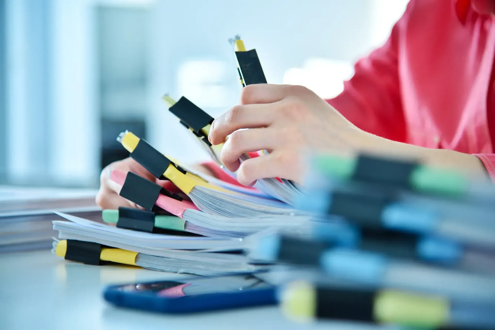 Manos organizan documentos en papel con clips de colores sobre un escritorio; hay un teléfono cerca.