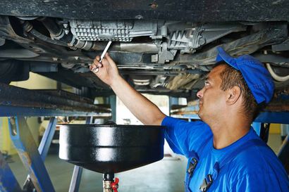 Un mécanicien en uniforme bleu travaille sous une voiture surélevée, en desserrant un bouchon de vidange d'huile au-dessus d'un entonnoir de récupération.