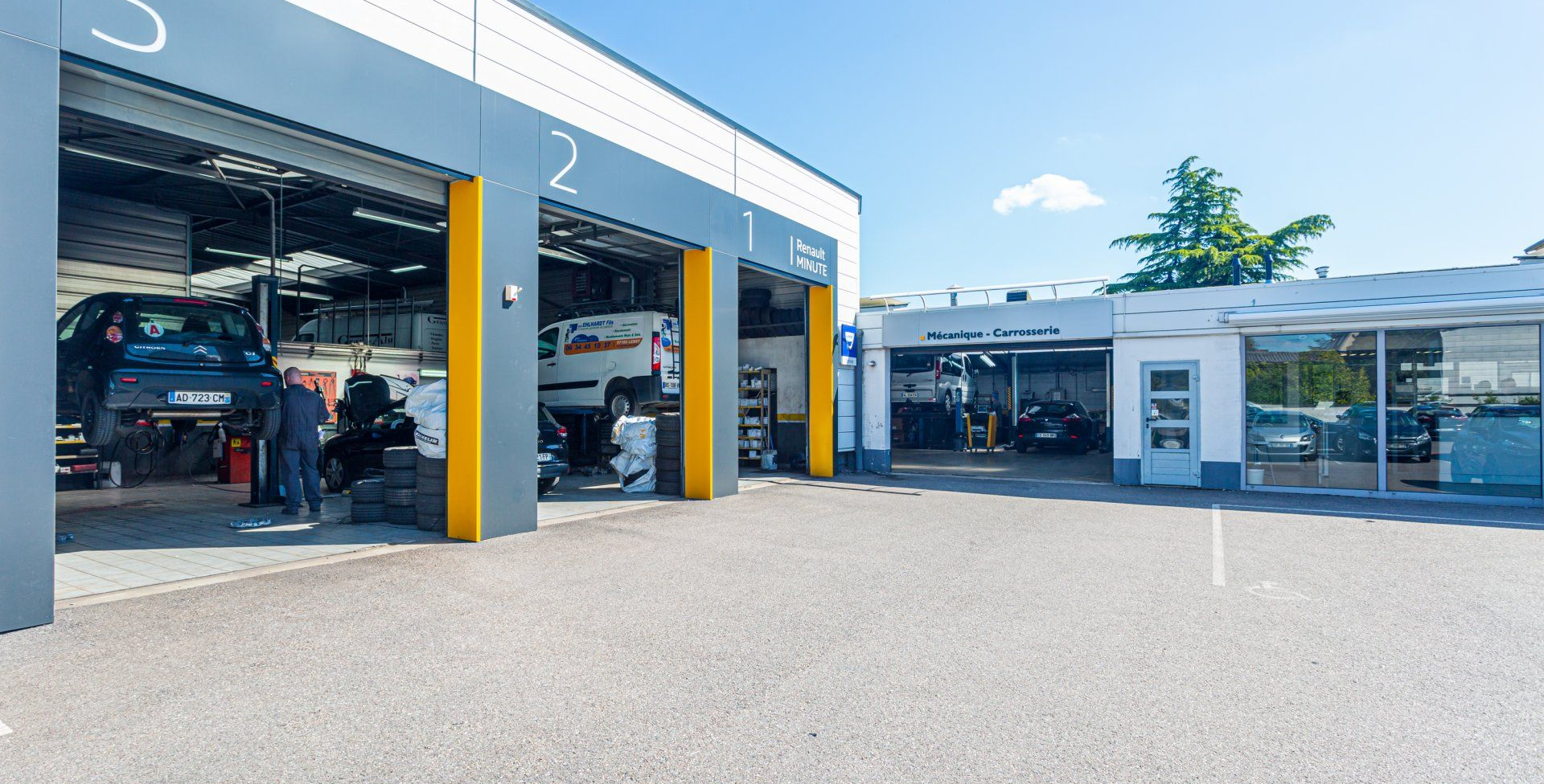 Extérieur d'un atelier de réparation automobile avec trois baies de service, des véhicules à l'intérieur et une entrée de bureau séparée au soleil.