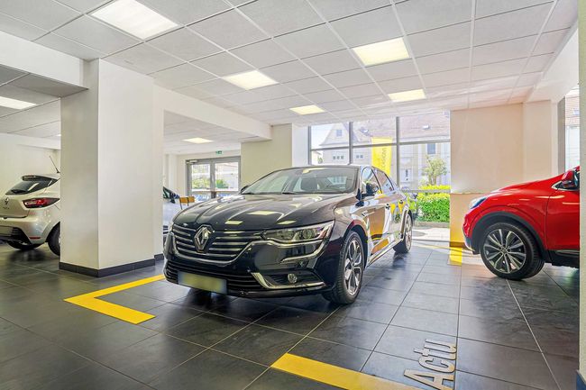 Un showroom moderne et lumineux chez un concessionnaire automobile, avec une berline Renault de couleur sombre au centre, flanquée d'autres voitures.
