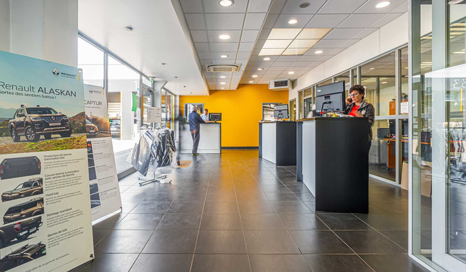 Un hall d'accueil du garage avec un mur jaune, un sol carrelé, des comptoirs d'accueil et un présentoir publicitaire automobile bien en vue.