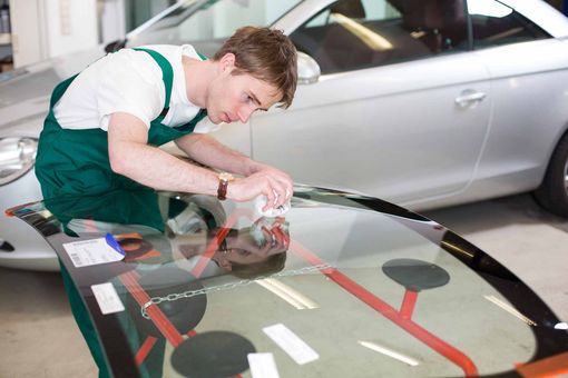 Un mécanicien en salopette verte nettoie le pare-brise d'une voiture dans un atelier de réparation automobile.