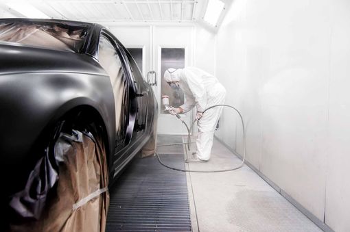 Une personne portant un équipement de protection utilise un pistolet à peinture pour peindre une voiture noire dans une cabine de peinture industrielle.