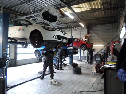 Des mécaniciens travaillent sur plusieurs voitures, dont une blanche et une rouge, à l'intérieur d'un atelier de réparation automobile lumineux.
