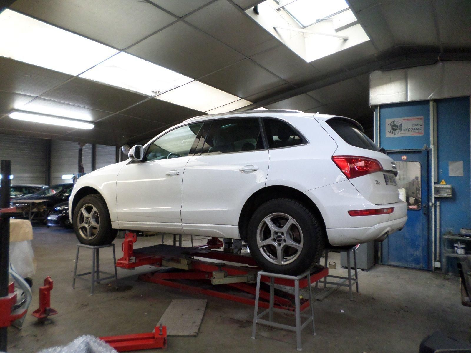 Un SUV blanc soulevé par un pont élévateur hydraulique rouge à l'intérieur d'un atelier de réparation automobile brillamment éclairé.