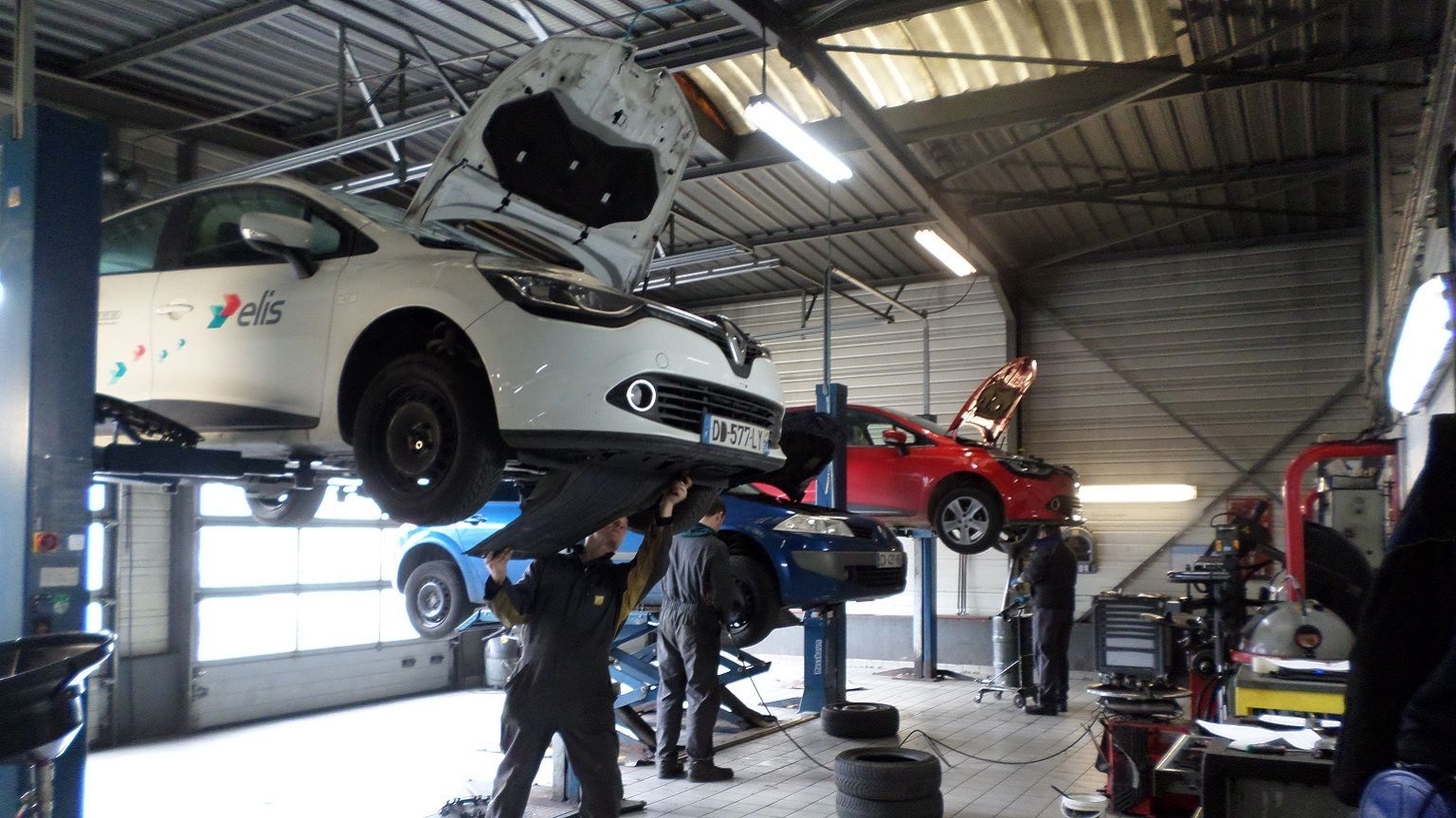Des mécaniciens travaillent sur des véhicules soulevés par des ponts élévateurs à l'intérieur d'un atelier de réparation automobile industriel et lumineux.