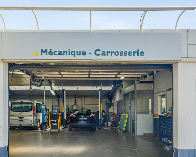 Un garage automobile ouvert avec une enseigne indiquant Mécanique - Carrosserie, montrant deux voitures à l'intérieur.