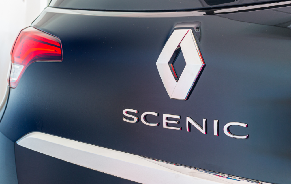 Gros plan sur l'arrière d'une Renault Scenic de couleur foncée, montrant le logo en forme de losange argenté et le mot SCENIC en chrome.