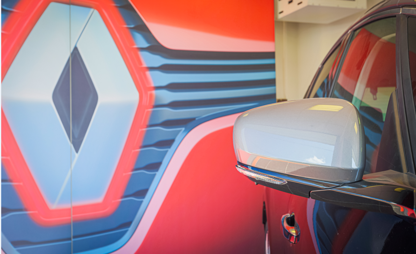 Rétroviseur latéral d'un véhicule garé devant un mur orné d'un grand logo Renault stylisé, rouge et bleu.