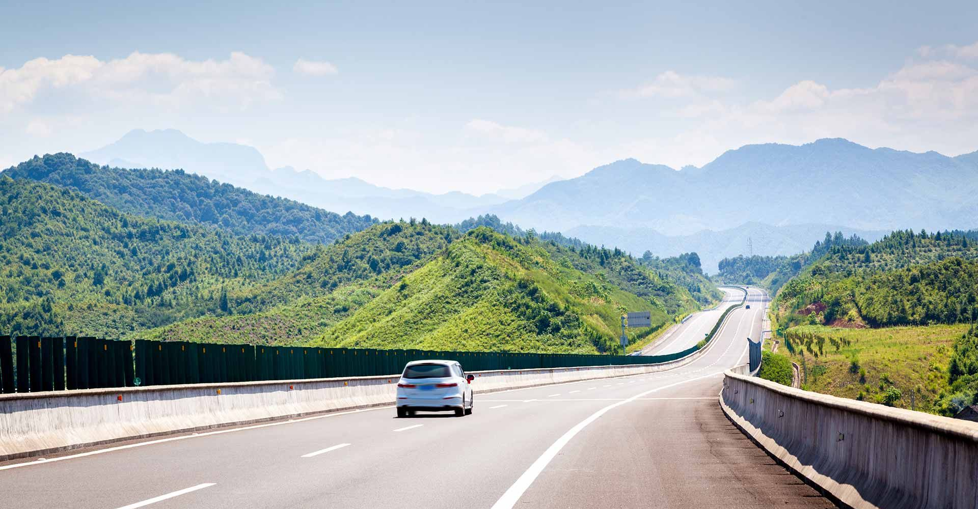 Une voiture blanche roule sur une autoroute dégagée, traversant un paysage montagneux verdoyant sous un ciel radieux et ensoleillé.