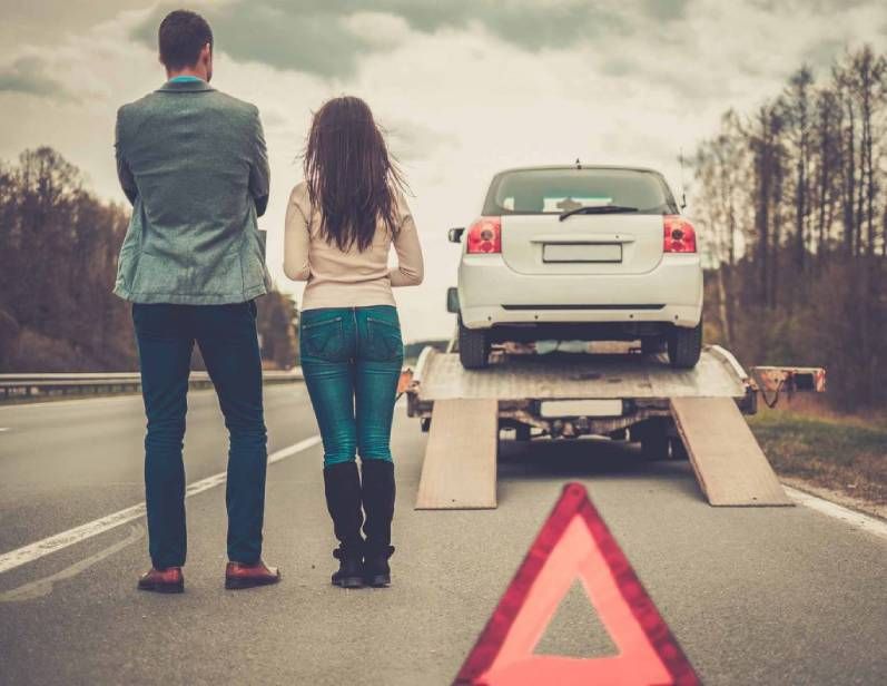 Un couple se tient sur une route à côté d'une voiture blanche qu'on charge sur une dépanneuse, avec un triangle de signalisation rouge au premier plan.