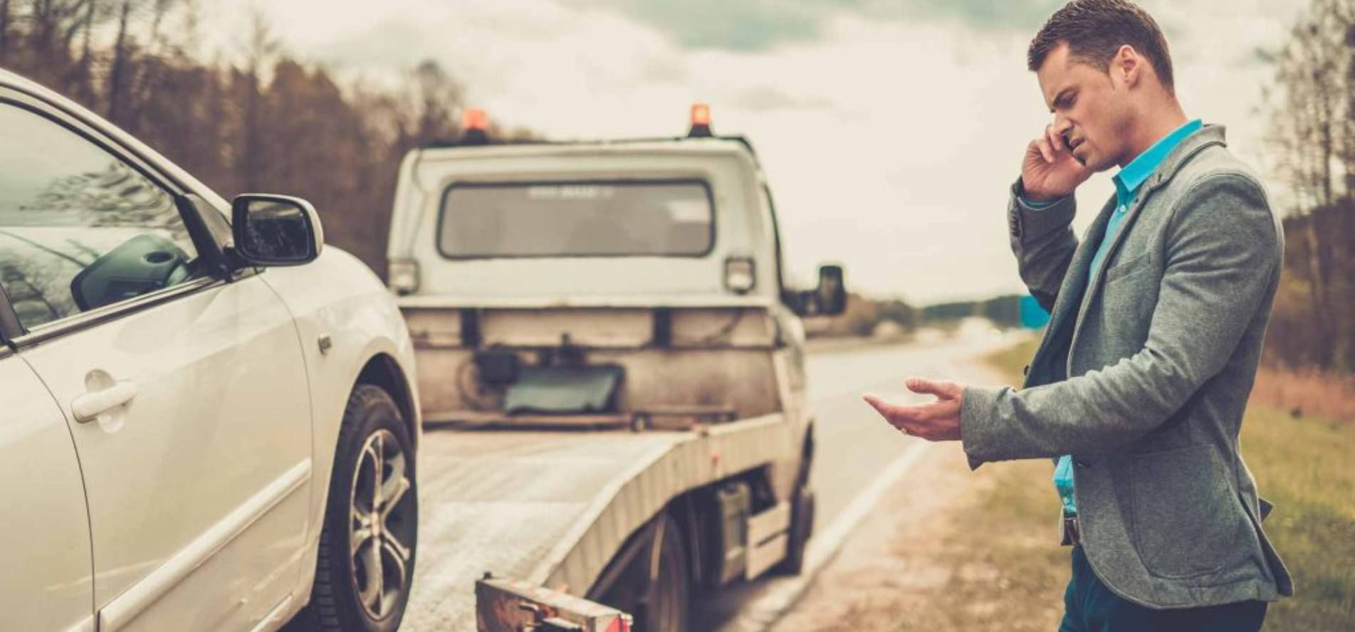 Une personne en tenue de ville parle au téléphone, debout au bord de la route, à côté de sa voiture qu'on charge sur une dépanneuse.