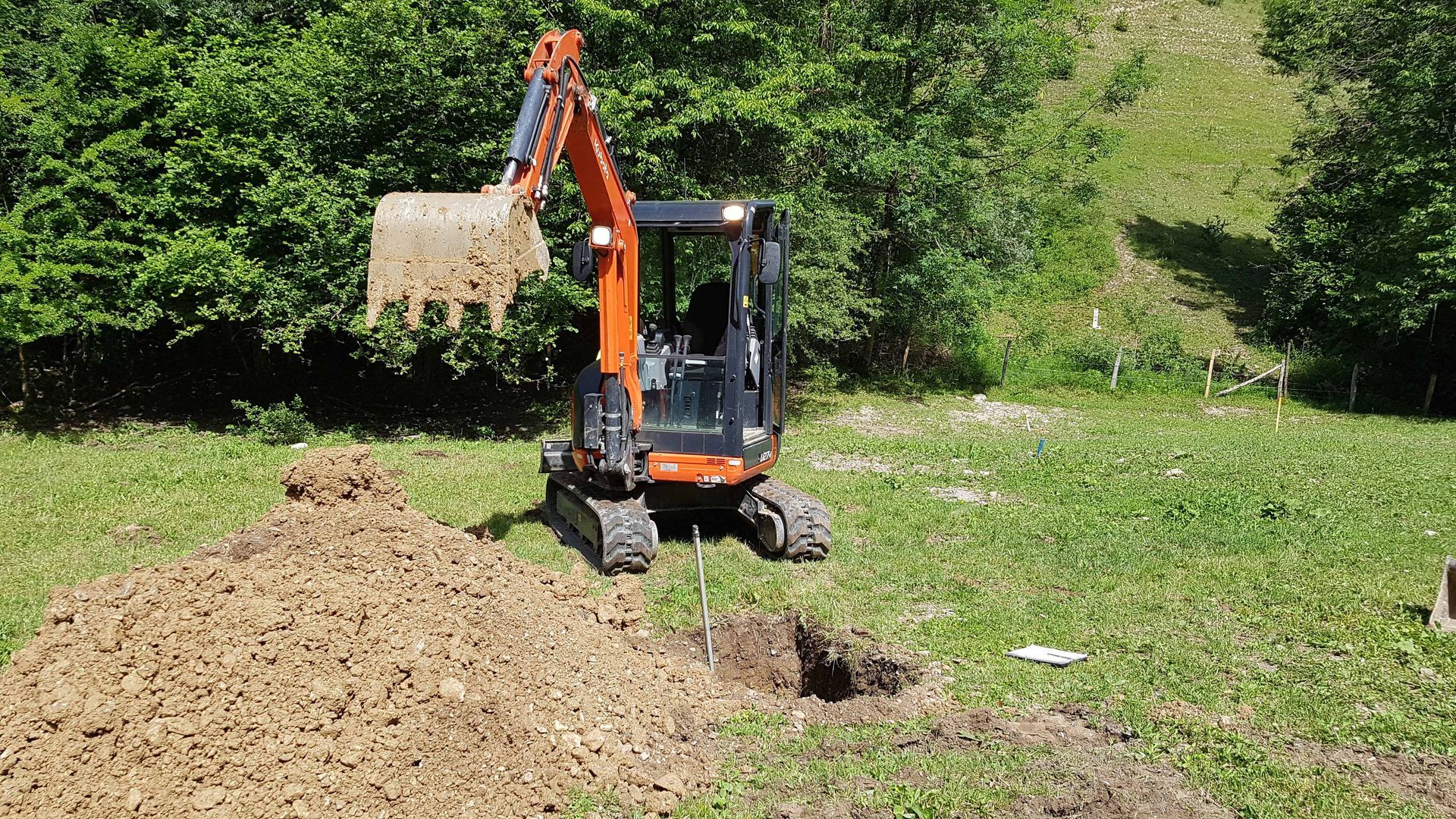 Une mini-pelle orange se dresse dans un champ herbeux, à côté d'un tas de terre excavée et d'un petit trou.