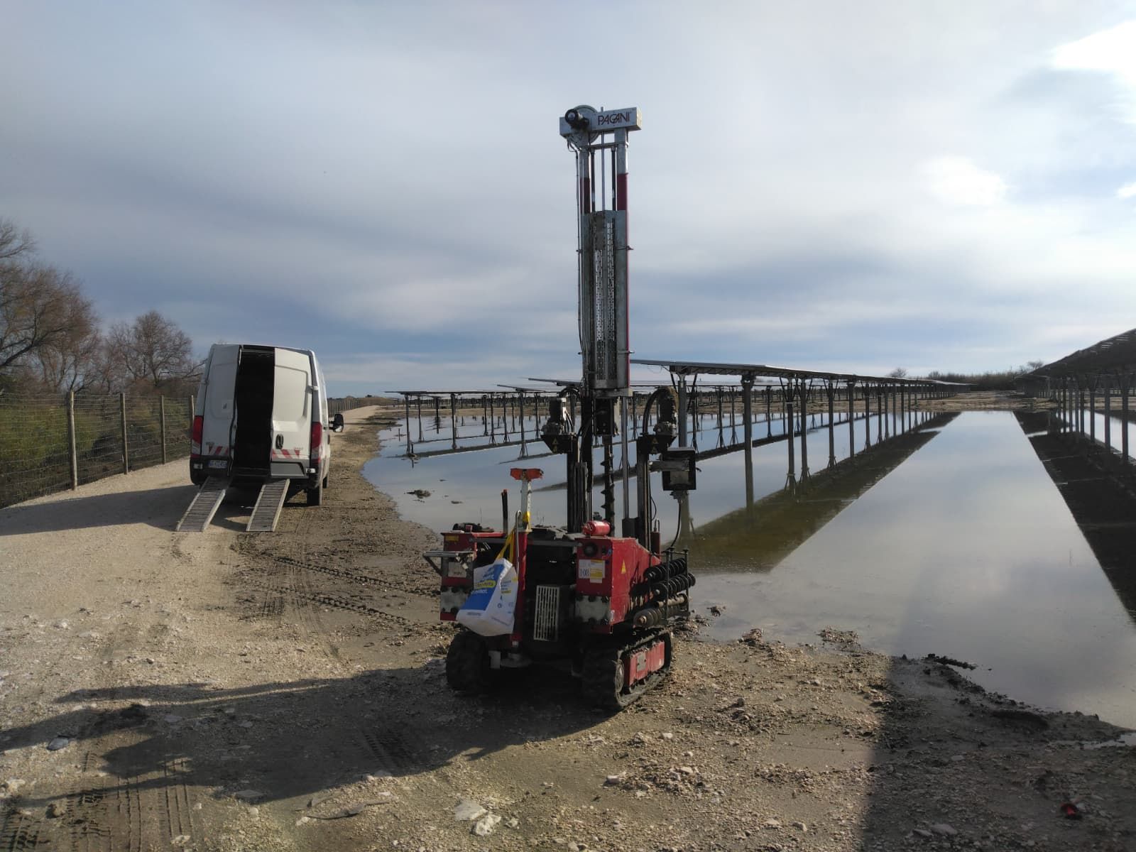 Une petite foreuse rouge à chenilles se dresse sur un chemin de terre à côté d'un champ de panneaux solaires inondé, avec une camionnette de chantier à proximité.