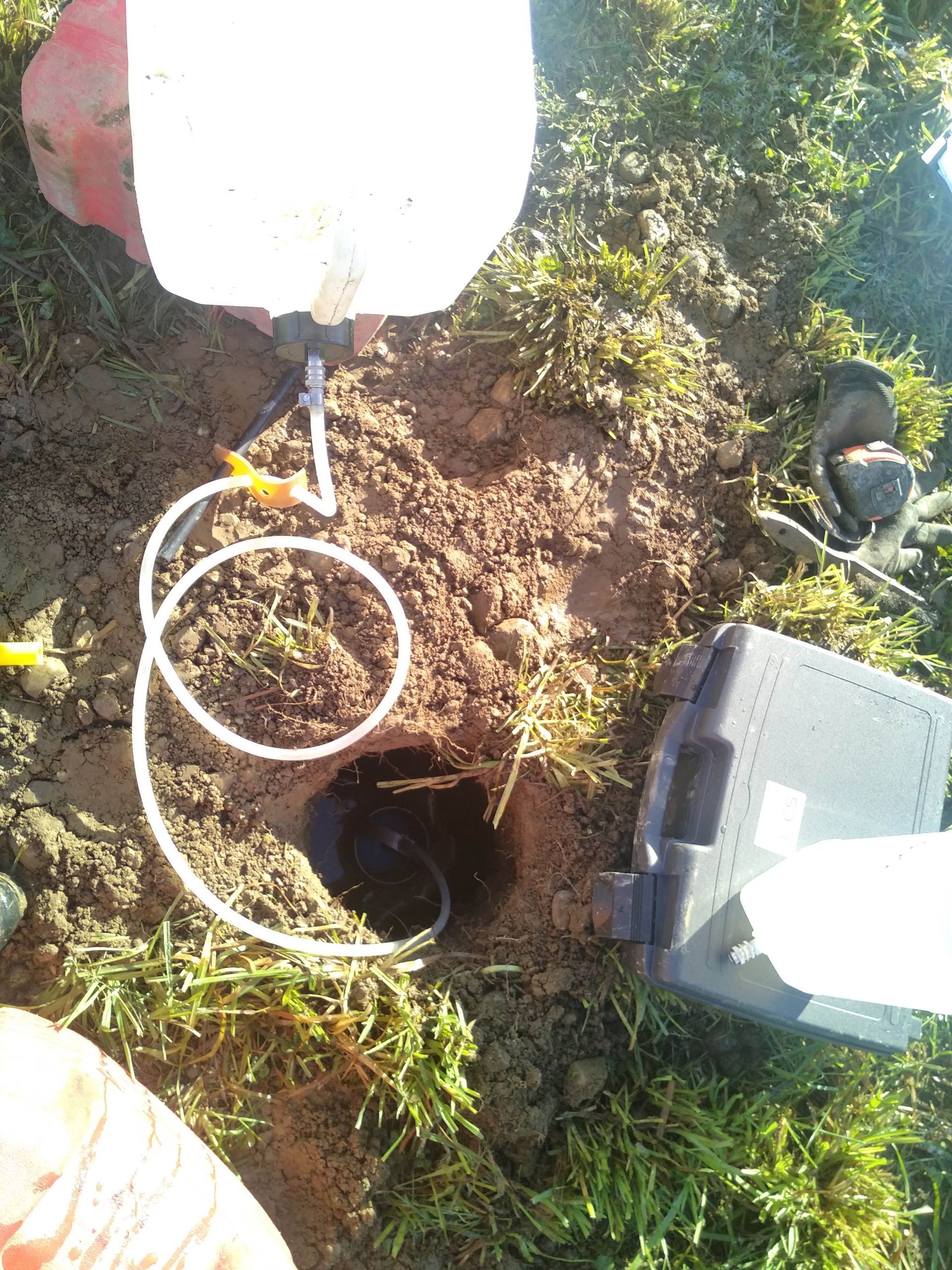 Un bidon blanc et une mallette noire pour le matériel sont posés sur l'herbe, avec des tubes plongeant dans un trou creusé dans la terre pour le prélèvement d'échantillons environnementaux.