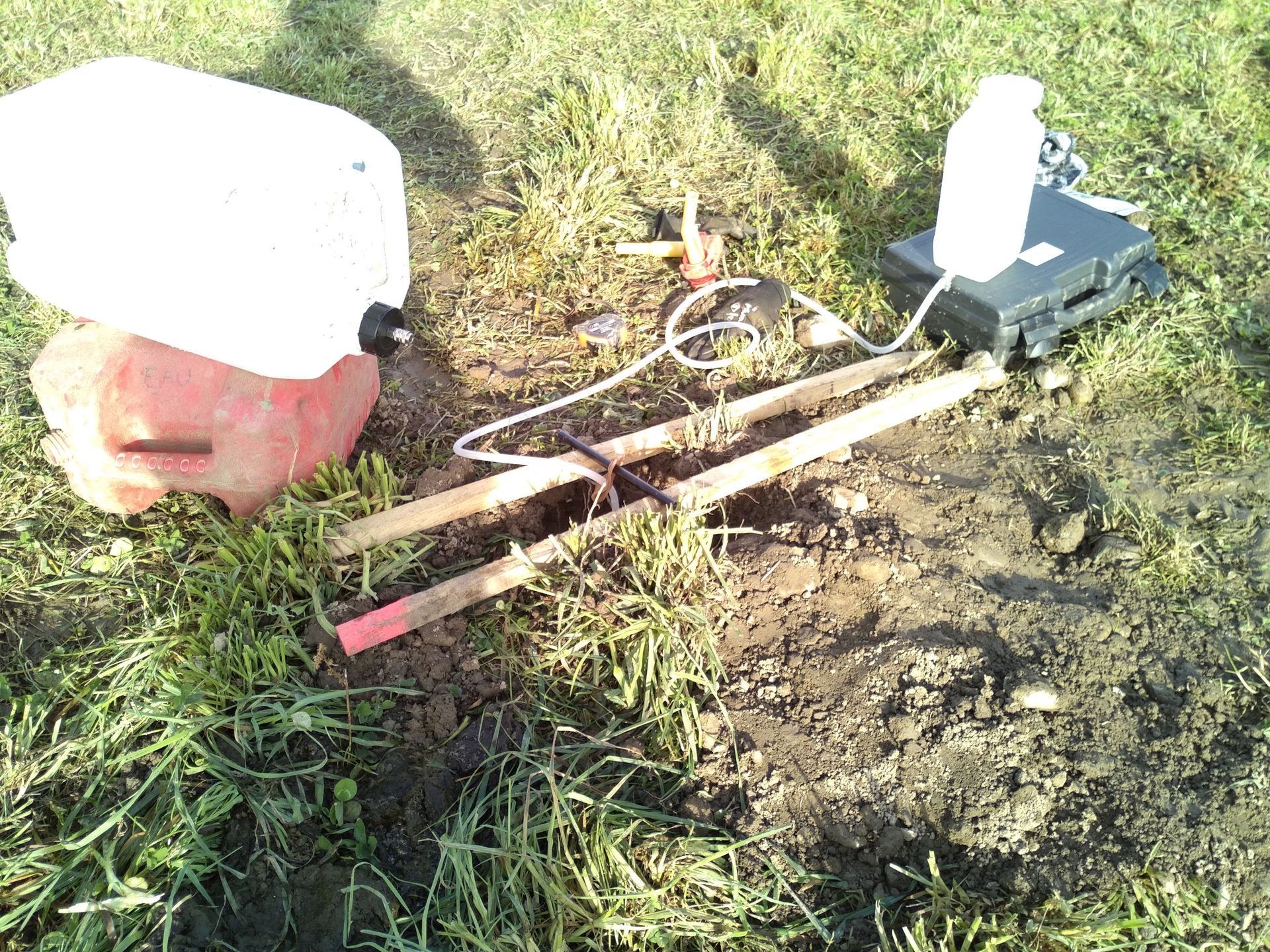 Le matériel de terrain pour les analyses de sol comprend un bidon en plastique, un écran noir et des piquets en bois reliés par des tubes sur l'herbe.