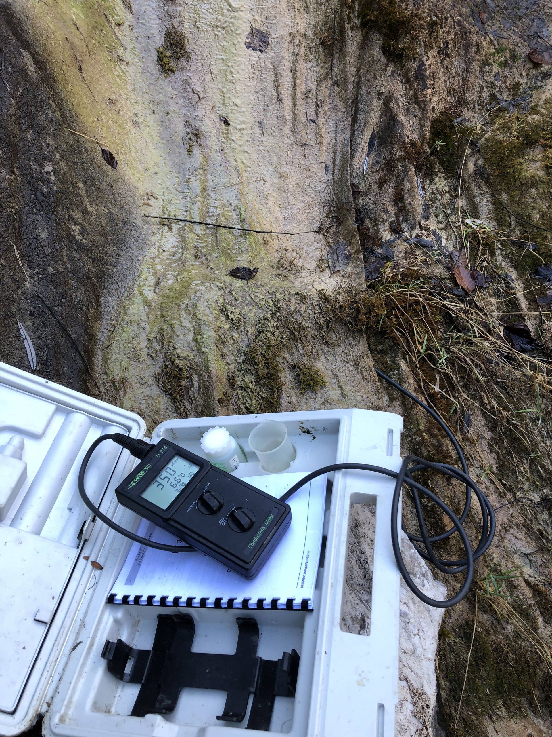 Une bouteille d'eau blanche reliée par un tuyau à un appareil noir et à un récipient cylindrique sombre, le tout dans une étroite tranchée de terre.