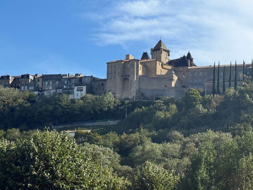 Château en pierre et village perchés sur une colline au-dessus d’arbres verdoyants, sous un ciel bleu dégagé.