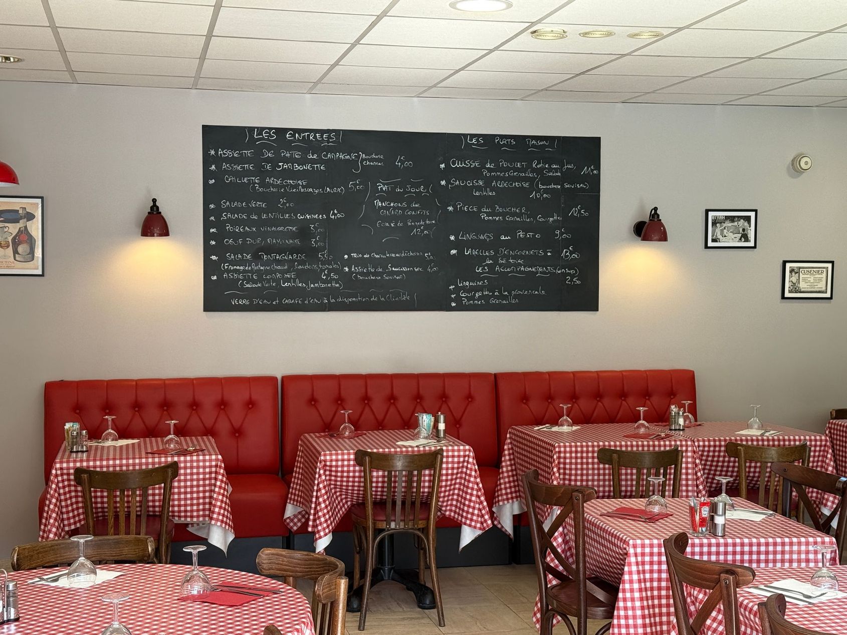 Café chaleureux avec banquettes rouges capitonnées, tables à carreaux et un grand tableau noir affichant le menu au mur.