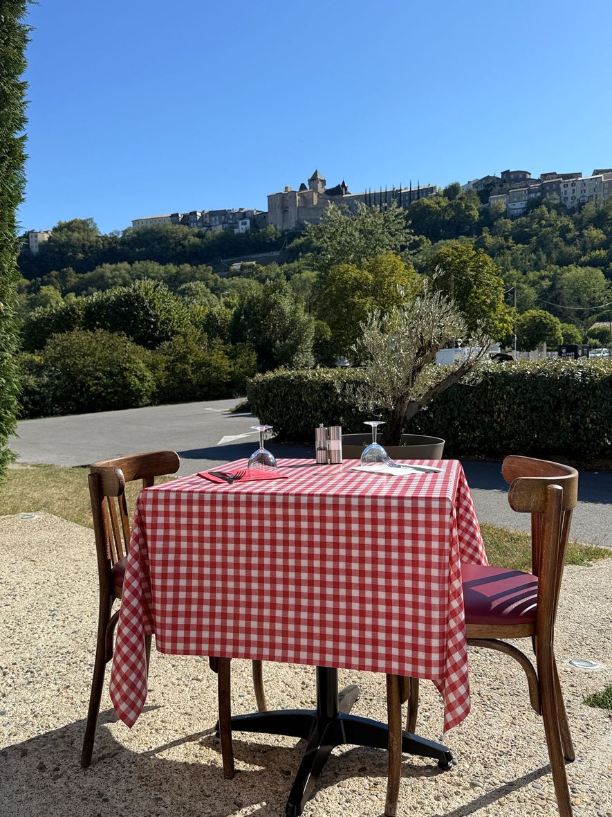 Table de café en plein air avec une nappe à carreaux rouges, deux chaises et un château perché sur une colline en arrière-plan