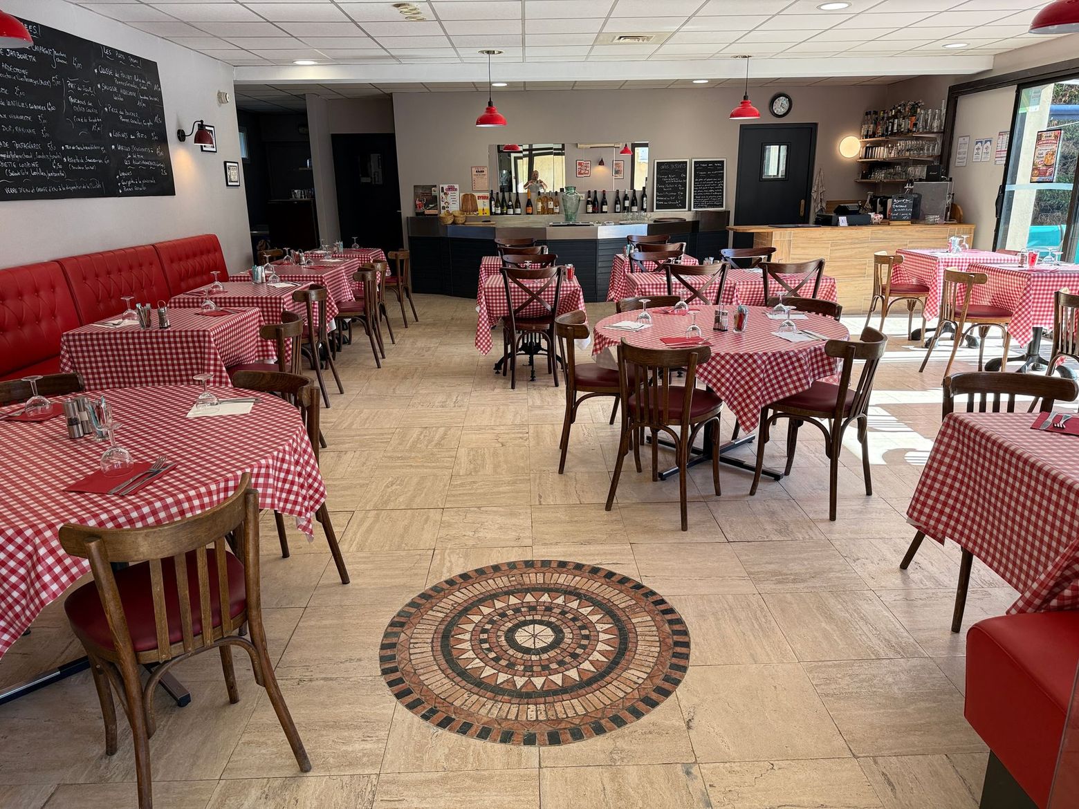 Salle à manger de restaurant vide, nappes à carreaux rouges, chaises en bois et mosaïque circulaire au sol.