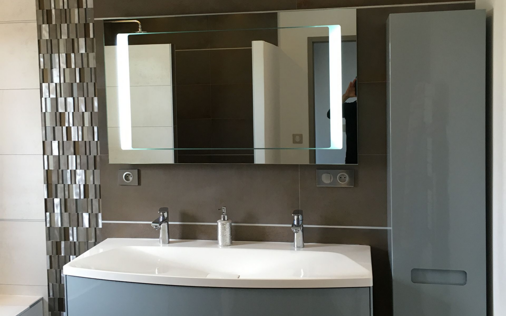 Salle de bains avec vasque grise, trois robinets, miroir éclairé et grande armoire de rangement.