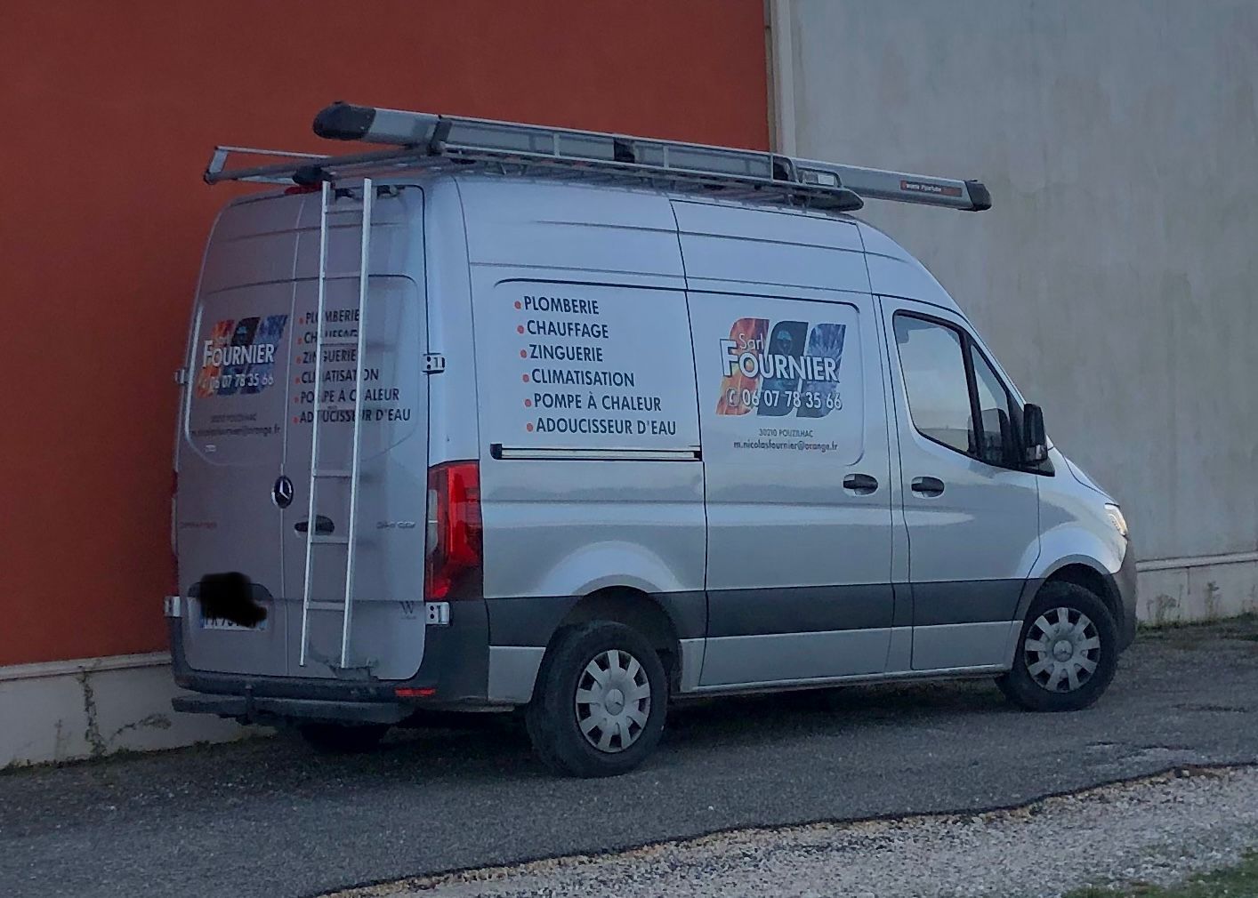 Fourgon de travail avec échelle sur le dessus, garé à côté d'un mur rouge.