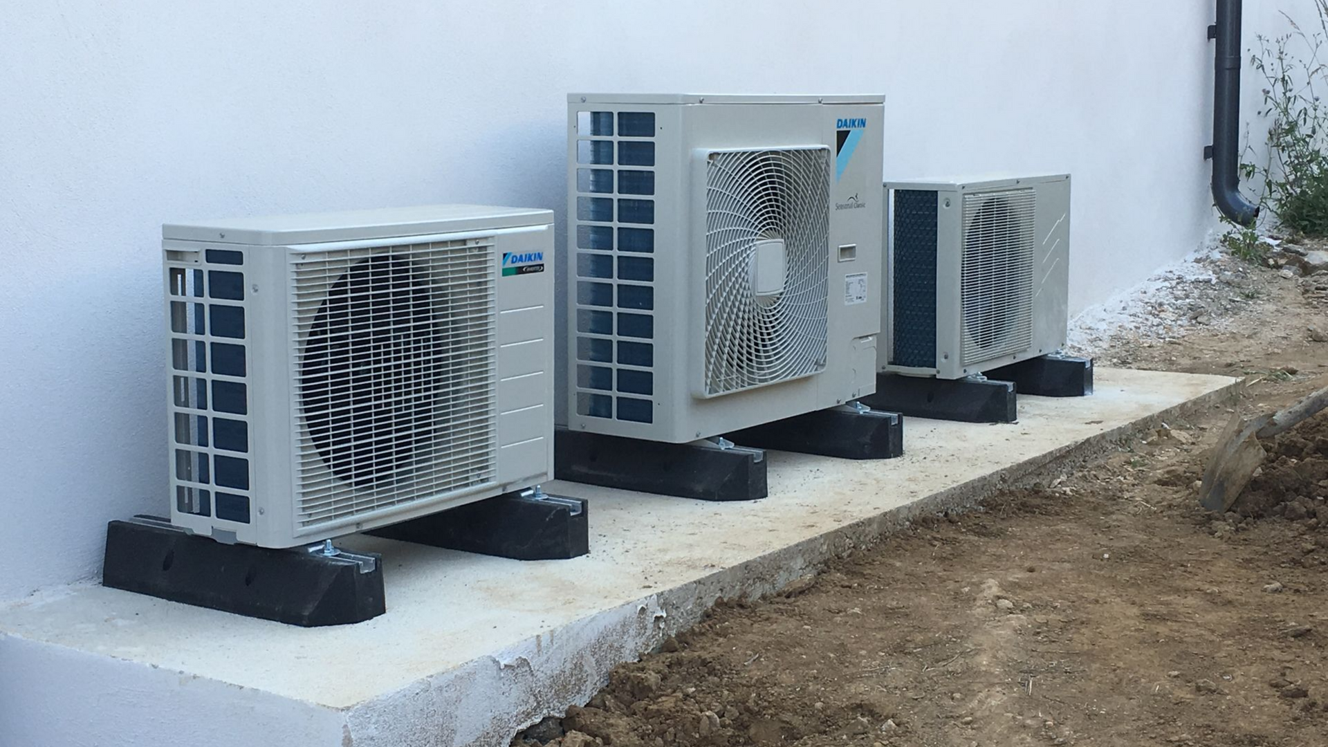 Trois unités de climatisation, montées sur des blocs noirs, sur une dalle de béton contre un mur blanc.