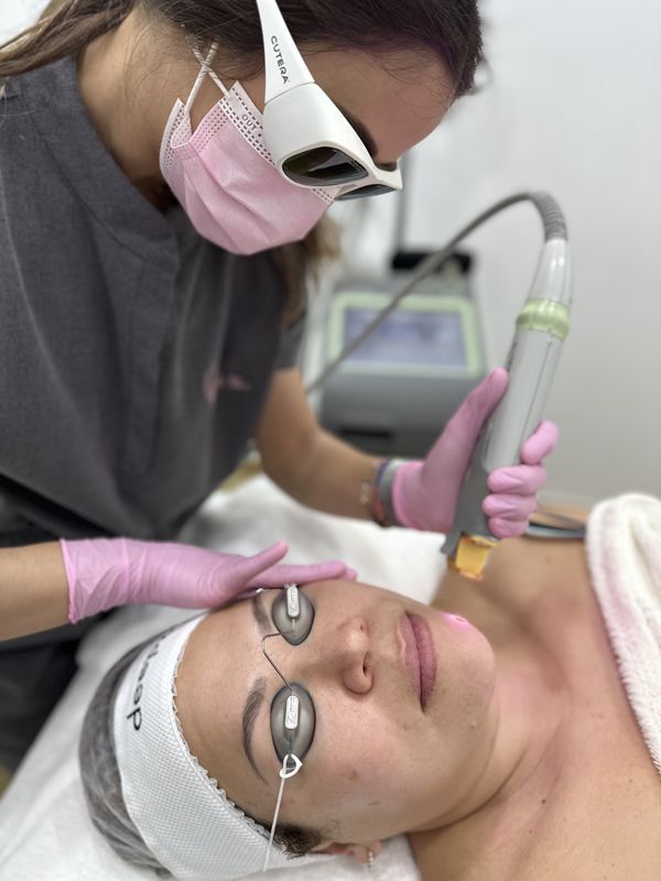 Persona recibiendo tratamiento láser facial en un entorno médico. La terapeuta, con guantes y mascarilla rosas, sostiene el dispositivo láser.