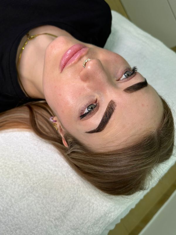 Mujer con cejas oscuras y angulosas sobre una superficie blanca, vestida con una camisa negra y un piercing en la nariz.