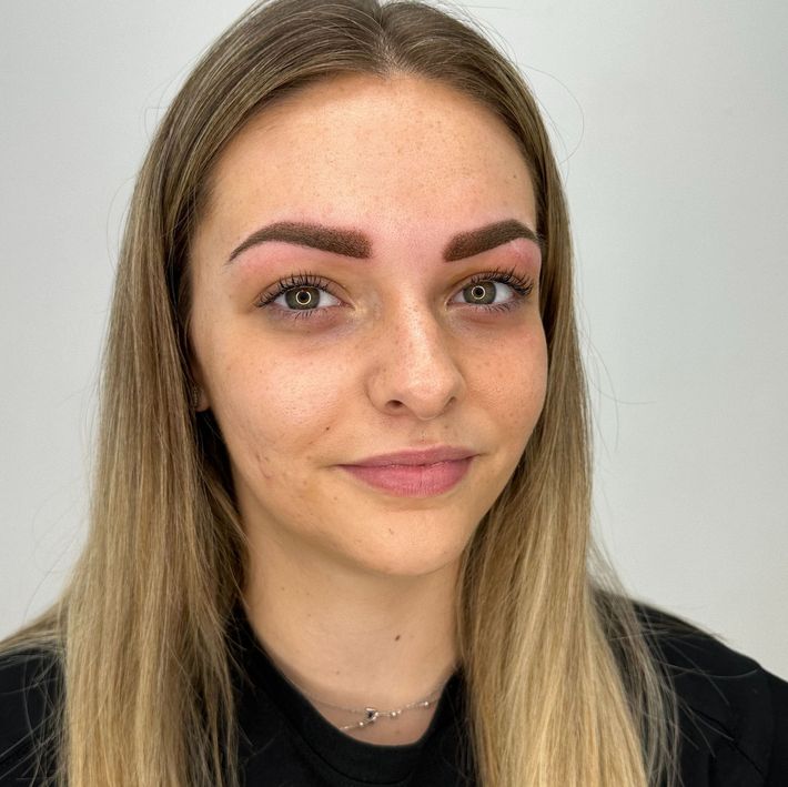 Mujer rubia con cejas recién tatuadas, sonriendo, vestida con una blusa negra.