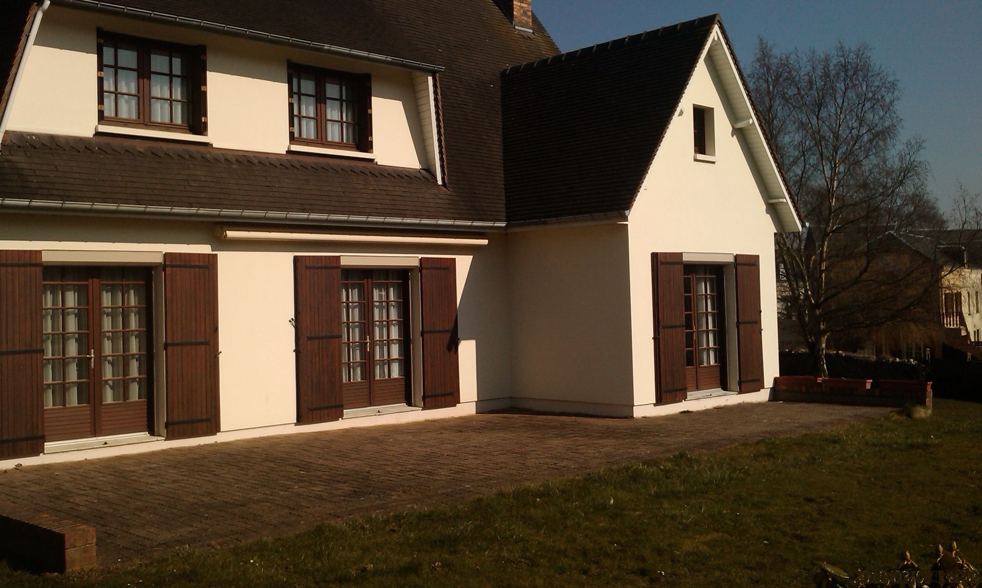 Une maison avec des murs beiges et des portes, fenêtres et volets marron