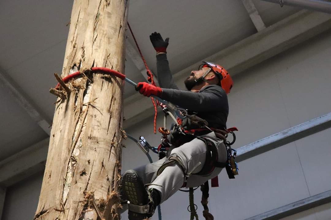 Hombre con equipo de seguridad trepando un poste de madera en un interior, intentando alcanzar algo hacia arriba.