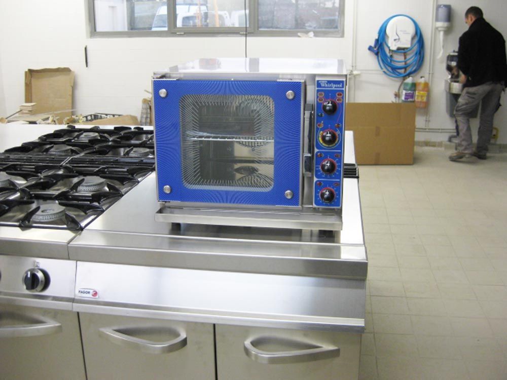 Four professionnel bleu et argent posé sur une plaque de cuisson en inox dans une cuisine. Une personne se tient près du mur.