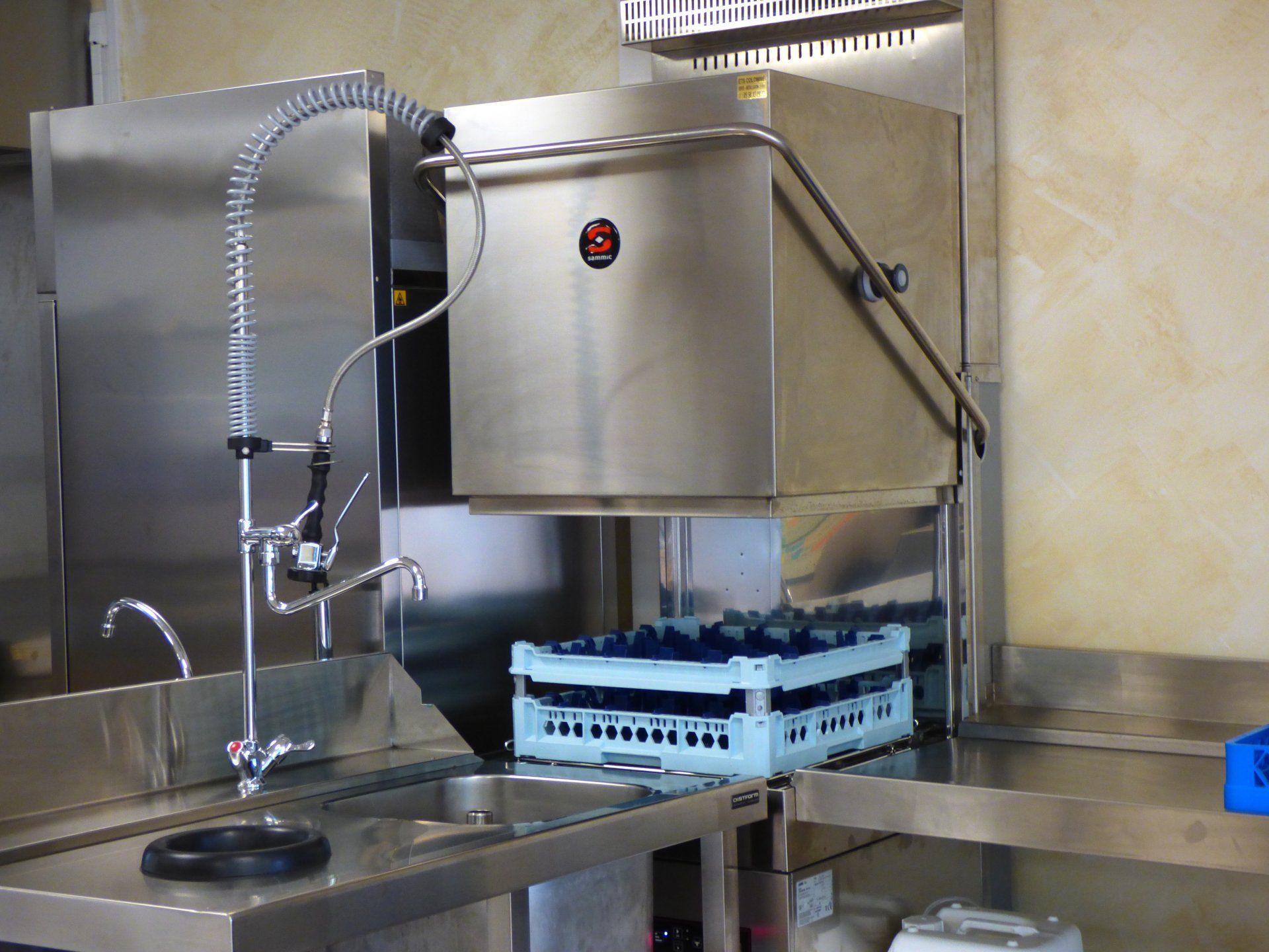 Lave-vaisselle professionnel dans une cuisine équipée d'un robinet à jet et d'un plateau à bouteilles.