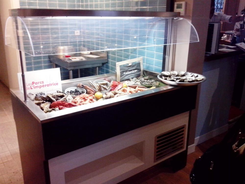 Vitrine de fruits de mer dans un restaurant, présentant divers coquillages et huîtres, sous un mur carrelé de bleu.