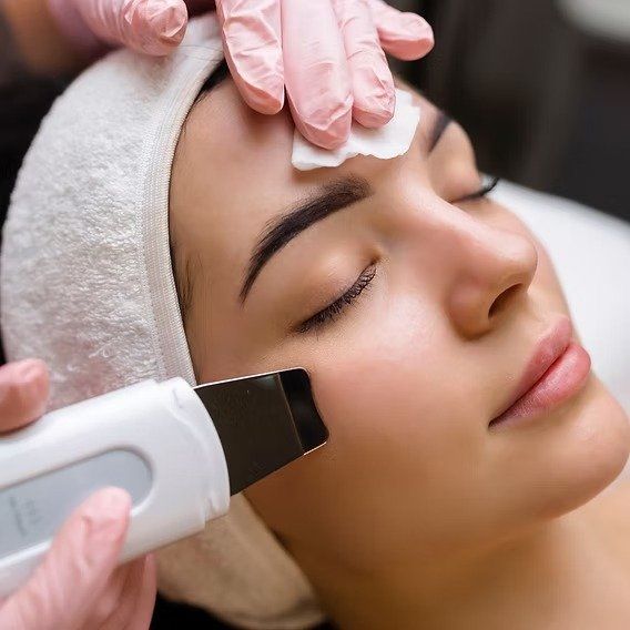Mujer recibiendo tratamiento facial; técnico utilizando depurador de piel ultrasónico, guantes rosas.